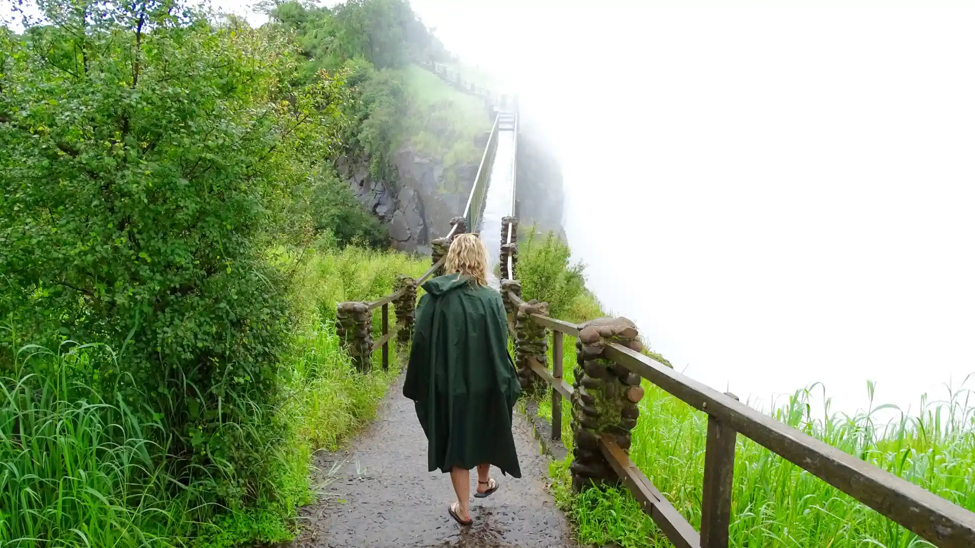 Lady walking along a path with a rail on opne side, bush on the left, Wearing a rain mac with water mist all around her