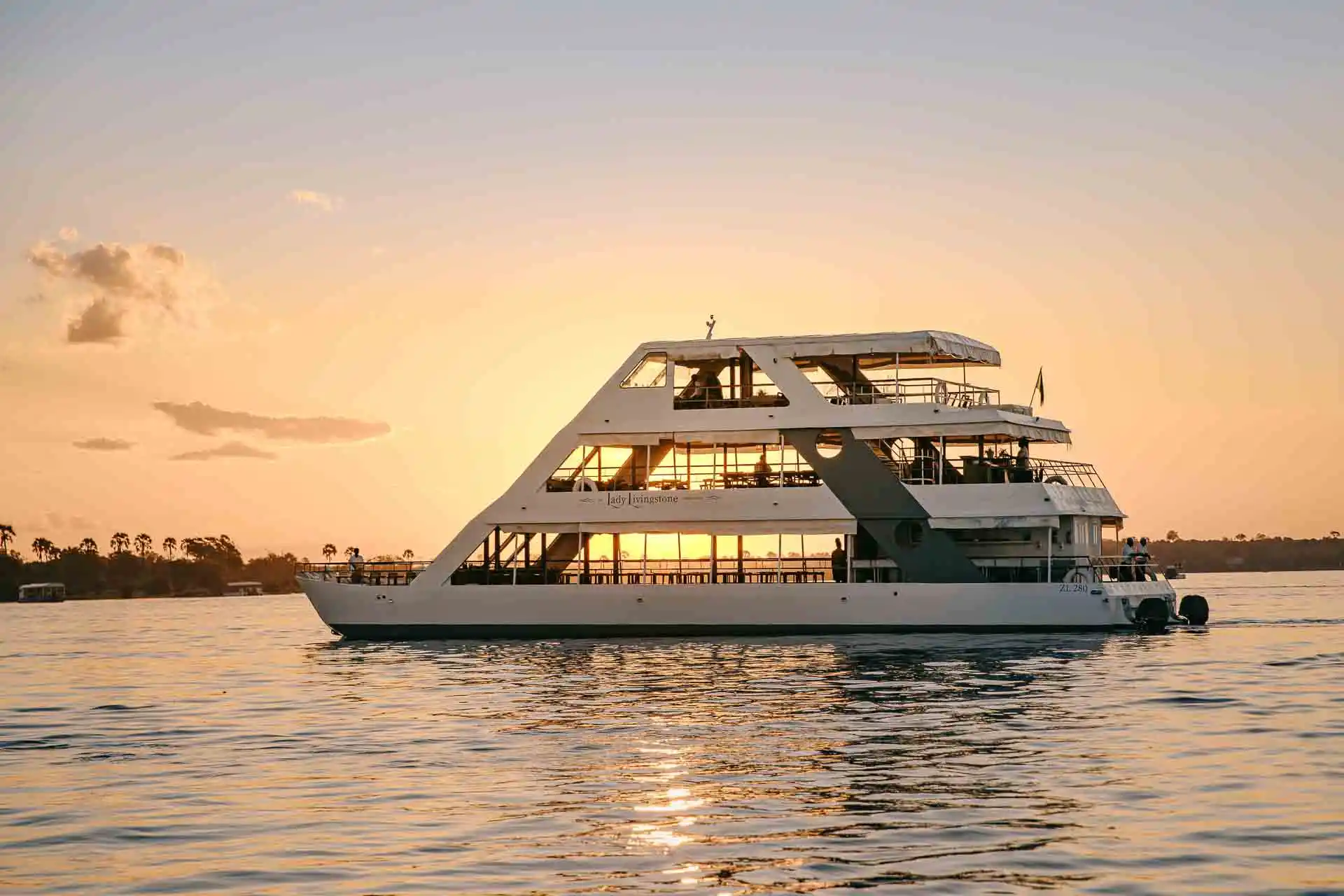 Floating on the water, the 3 decks of the lady livingstone visible with sunset shining through. Water all around and the river bank in the background