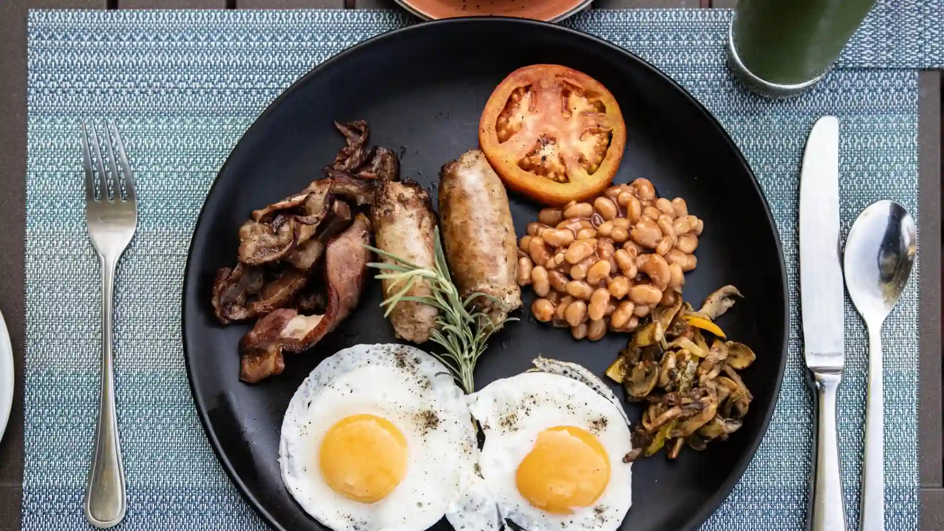 A dark breakfast plate with sausages, beans, bacon, eggs and mushrooms on it